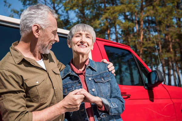 Sonriendo pareja mayor de turistas de pie cerca de coche rojo, tomados de la mano y abrazándose en el día soleado - foto de stock