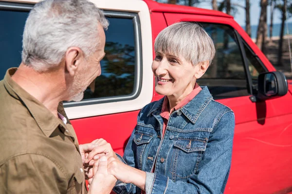 Souriant couple de touristes âgés debout près de voiture rouge, se tenant la main et embrassant dans la journée ensoleillée — Photo de stock