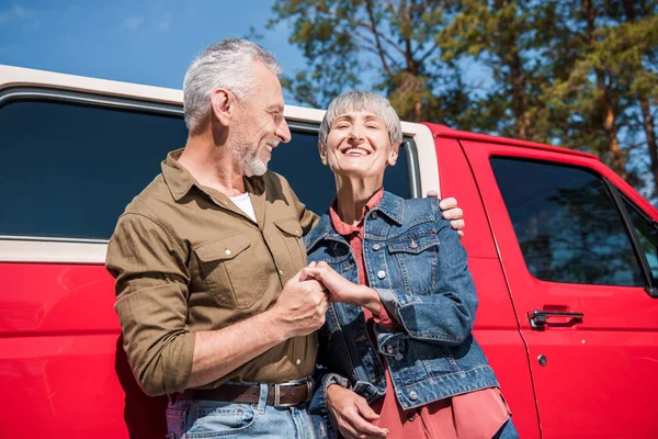 Sonriendo pareja mayor de turistas de pie cerca de coche rojo, tomados de la mano y abrazándose en el día soleado - foto de stock