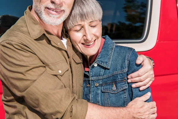 Cropped view of smiling senior couple of tourists standing near red car and embracing in sunny day — Stock Photo