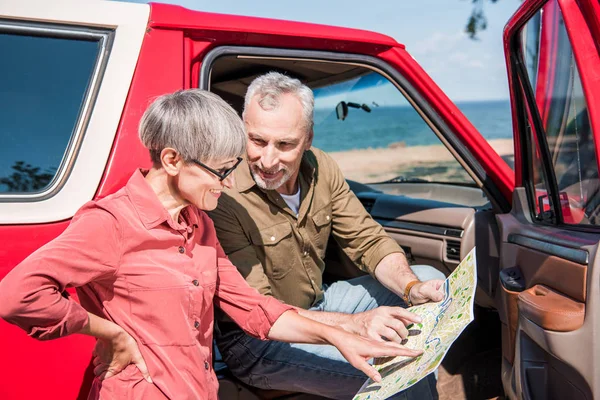 Усміхнений старший мандрівник сидить у червоній машині і дивиться на карту з дружиною — стокове фото
