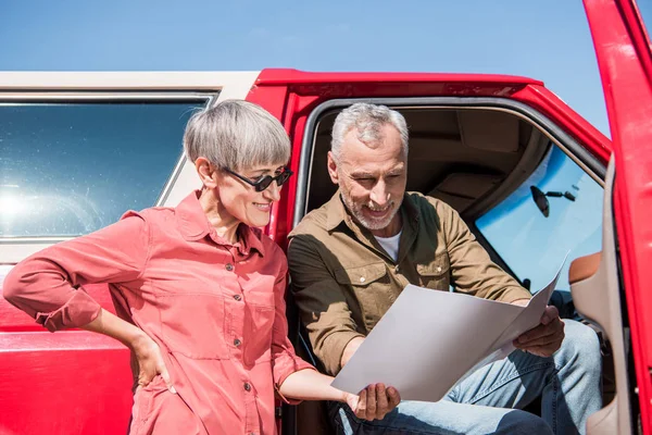 Sorrindo viajante sênior sentado em carro vermelho e olhando para o mapa com a esposa — Fotografia de Stock