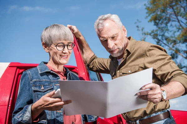 Senior couple of travelers standing near car and looking at map in sunny day — Stock Photo