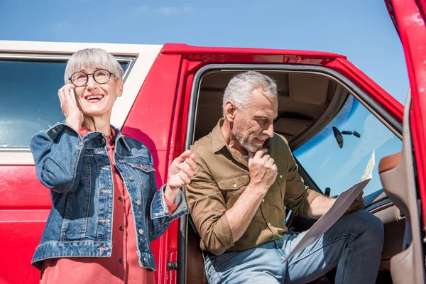 Senior woman in glasses talking on smartphone while husband sitting in car and looking at map — Stock Photo