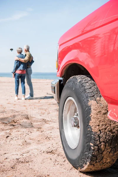 Visão de comprimento total do casal sênior abraçando e tomando selfie com carro vermelho em primeiro plano — Fotografia de Stock