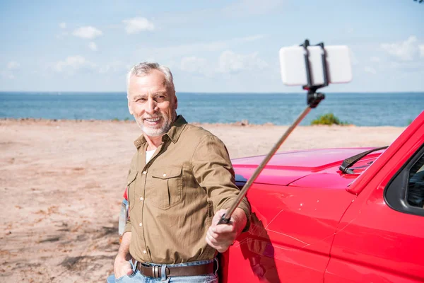 Улыбающийся пожилой человек, стоящий возле красной машины и делающий селфи в солнечный день — стоковое фото