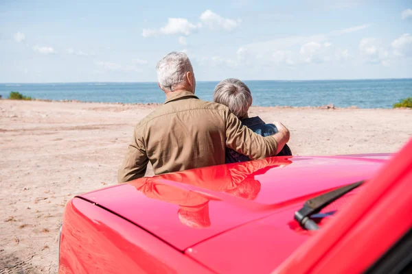 Back view of senior couple standing near red car at beach and embracing in sunny day — Stock Photo