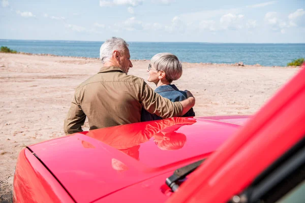 Vue arrière du couple de personnes âgées debout près de la voiture rouge à la plage et embrassant dans la journée ensoleillée — Photo de stock