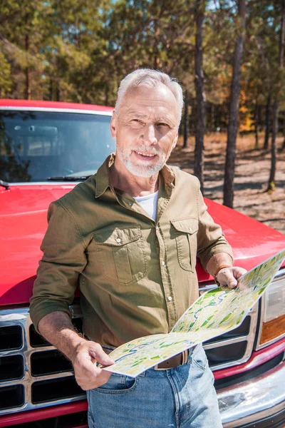 Sorrindo turista sênior em pé perto de carro vermelho e segurando mapa em dia ensolarado — Fotografia de Stock