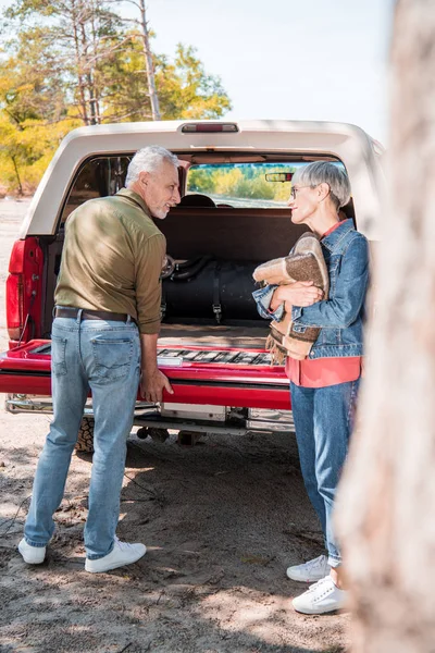 Full length view of senior couple of tourists with blanket standing near car and looking at each other — Stock Photo