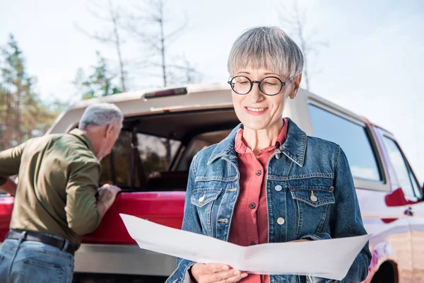 Souriant femme âgée tenant carte tandis que le mari debout près de la voiture dans la journée ensoleillée — Photo de stock