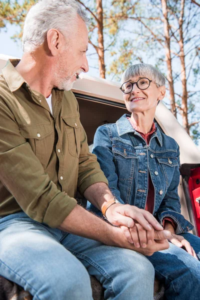 Pareja de ancianos en jeans tomados de la mano y mirándose con sonrisa - foto de stock