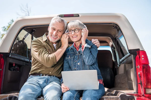 Smiling senior couple of tourists with laptop listening music in headphones in car — Stock Photo
