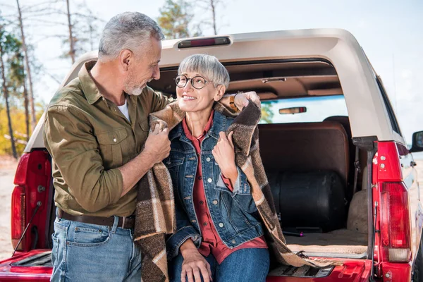 Sonriente pareja de ancianos sentados en el coche con manta y mirándose - foto de stock