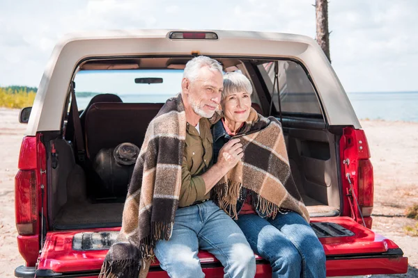 Sonriente pareja de ancianos sentado en el coche con manta - foto de stock