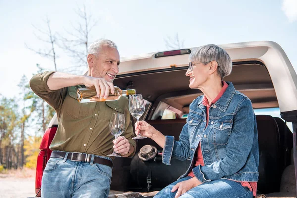 Souriant homme âgé verser du vin dans des verres à vin et regarder femme près de la voiture — Photo de stock