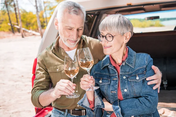 Glückliches Senioren-Paar klimpert mit Weingläsern und umarmt nahe Auto — Stockfoto