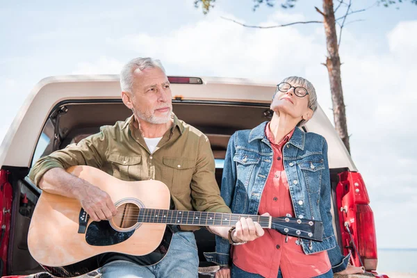 Senior man playing acoustic guitar while standing near car with wife — Stock Photo