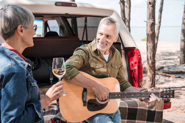 Senior man playing acoustic guitar while standing near car with wife — Stock Photo