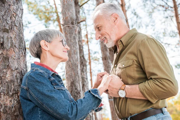 Happy senior couple holding hands and looking at each other near car in forest — Stock Photo