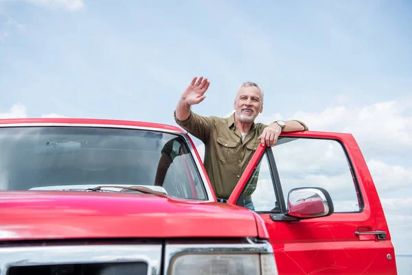 Senior Mann im Hemd in rotem Auto winkt Hand und lächelt an sonnigem Tag — Stockfoto