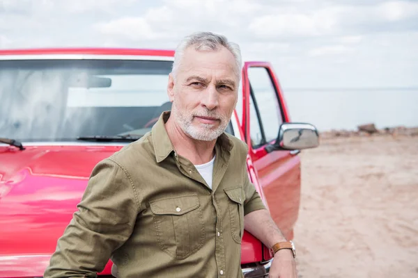 Uomo anziano in camicia in piedi vicino auto rossa in spiaggia — Foto stock