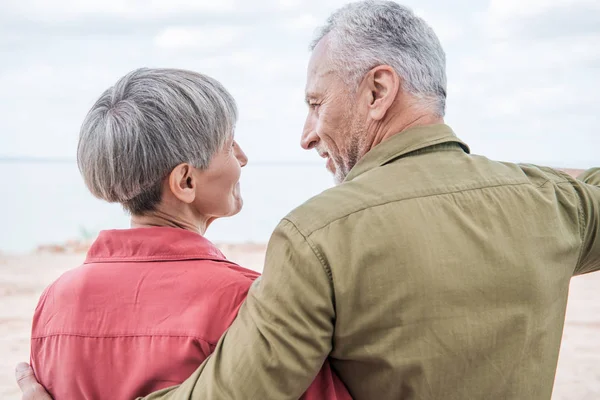 Rückansicht eines älteren Paares, das sich am Strand umarmt und ansieht — Stockfoto