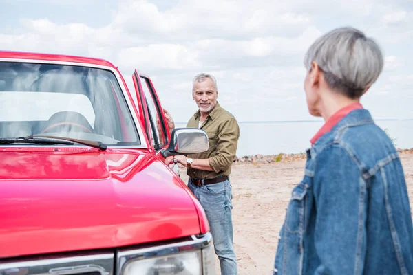 Selective focus of senior couple of tourists standing near red car and looking at each other at beach — Stock Photo