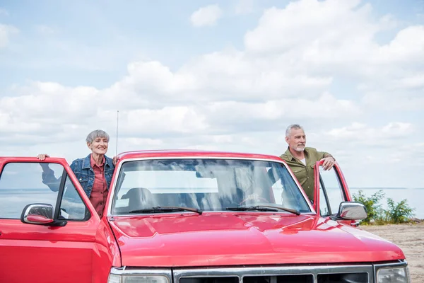 Pareja de turistas mayores de pie cerca de coche rojo en la playa - foto de stock