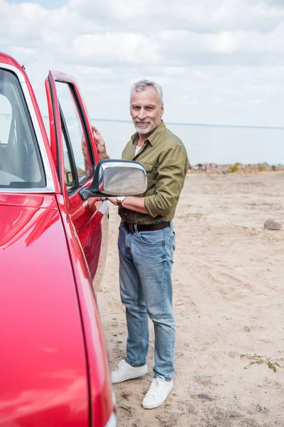 Pleine longueur vue de sourire homme âgé debout près de voiture rouge à la plage — Photo de stock