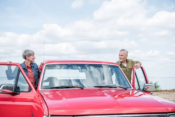 Усміхнена старша пара туристів стоїть біля червоної машини і дивиться один на одного на пляжі — стокове фото
