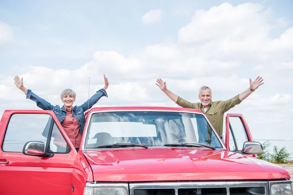 Усміхнена старша пара туристів стоїть з руками вгору біля червоної машини на пляжі — стокове фото
