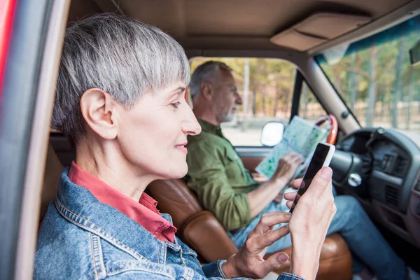 Seitenansicht einer lächelnden Seniorin mit Smartphone mit leerem Bildschirm, während ihr Mann im Auto auf die Landkarte schaut — Stockfoto