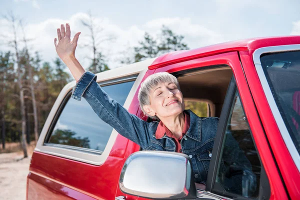 Mulher sênior feliz acenando mão da janela do carro com os olhos fechados — Fotografia de Stock