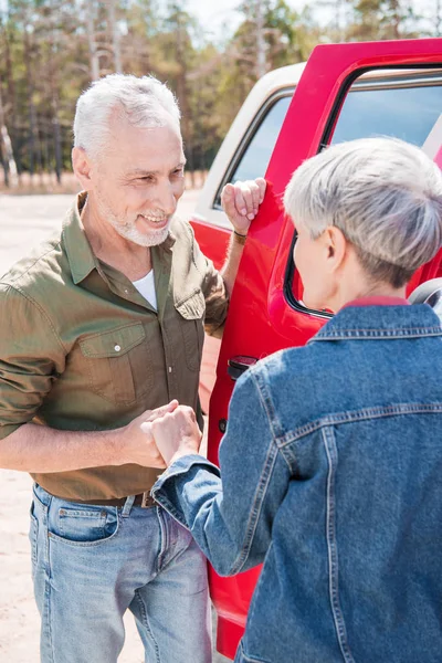 Couple âgé souriant debout près de voiture rouge, se tenant la main et se regardant — Photo de stock