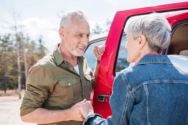 Sonriente pareja de ancianos de pie cerca de coche rojo, tomados de la mano y mirándose - foto de stock