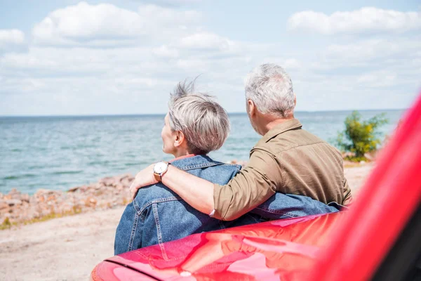 Задний вид пожилой пары, стоящей рядом с красной машиной и обнимающейся на пляже — стоковое фото