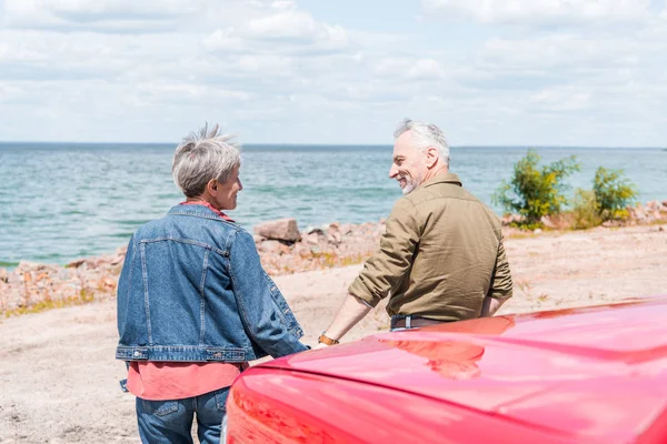 Vista trasera de la pareja de ancianos de pie cerca de coche rojo y mirándose en la playa - foto de stock