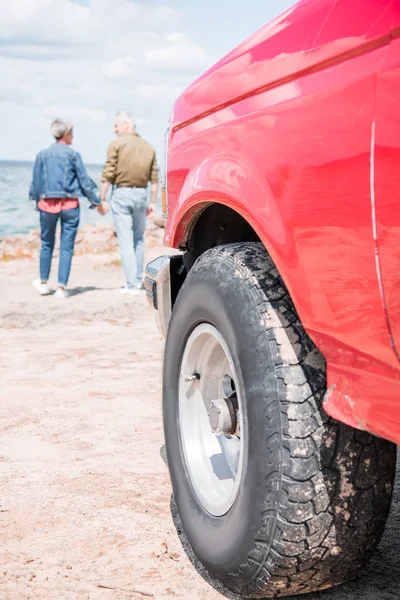 Full length view of senior couple holding hands while walking at beach and red car on foreground — Stock Photo