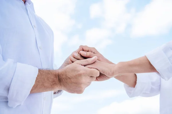 Vista recortada de pareja mayor en camisas blancas cogidas de la mano bajo el cielo azul - foto de stock