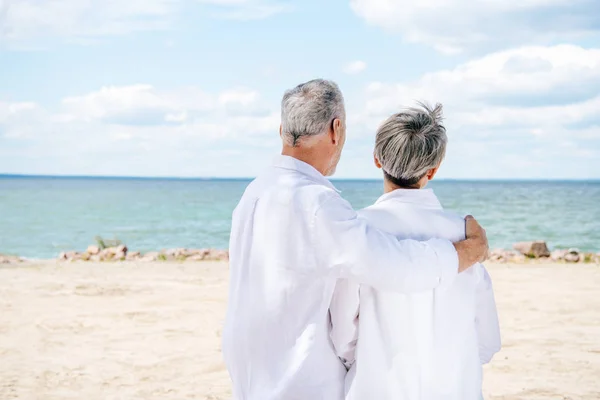 Visão traseira do casal sênior em camisas brancas abraçando na praia — Fotografia de Stock