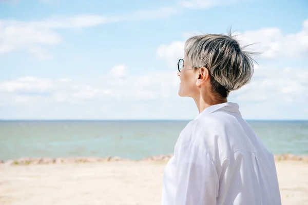 Anziana donna in camicia bianca guardando lontano dalla spiaggia — Foto stock