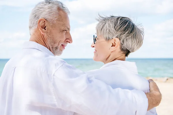 Sonriente pareja de ancianos en camisas blancas abrazándose y mirándose cerca del río - foto de stock