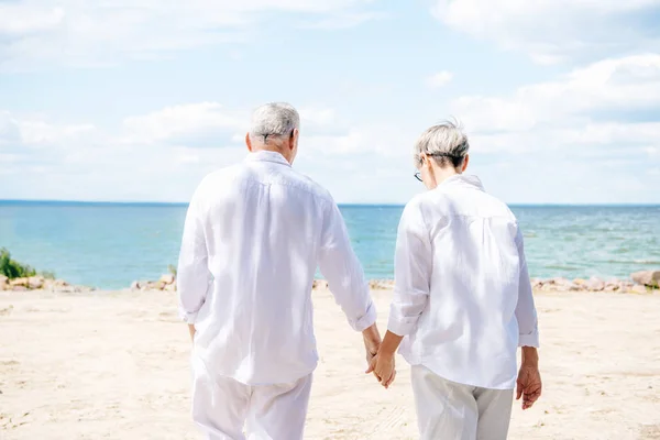 Vista trasera de pareja mayor en camisas blancas cogidas de la mano en la playa - foto de stock