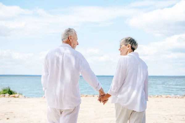 Visão traseira do casal sênior em camisas brancas de mãos dadas e olhando uns para os outros na praia — Fotografia de Stock