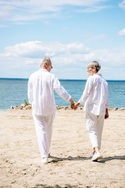 Back view of senior couple in white shirts holding hands and looking at each other at beach — Stock Photo