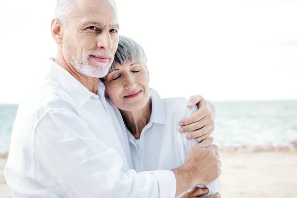 Feliz pareja de ancianos en camisas blancas abrazando en la playa - foto de stock