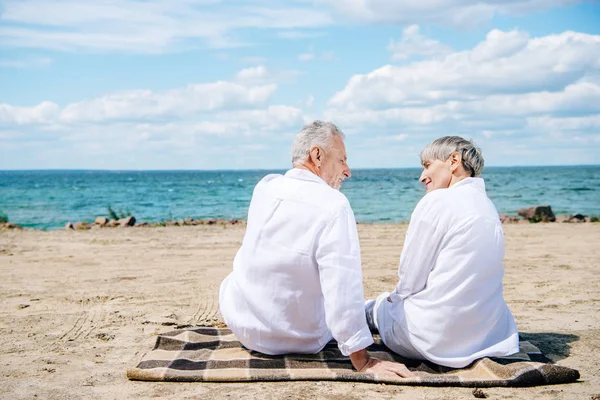 Rückansicht eines älteren Paares in weißen Hemden, das auf einer Decke sitzt und sich am Strand anschaut — Stockfoto