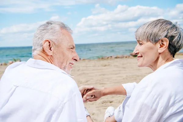 Sonriente pareja de ancianos tomados de la mano y mirándose en la playa - foto de stock