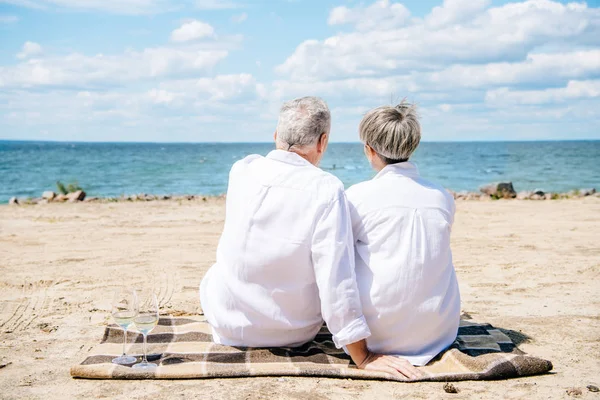 Rückansicht eines älteren Ehepaares in weißen Hemden, das auf einer Decke am Strand sitzt — Stockfoto